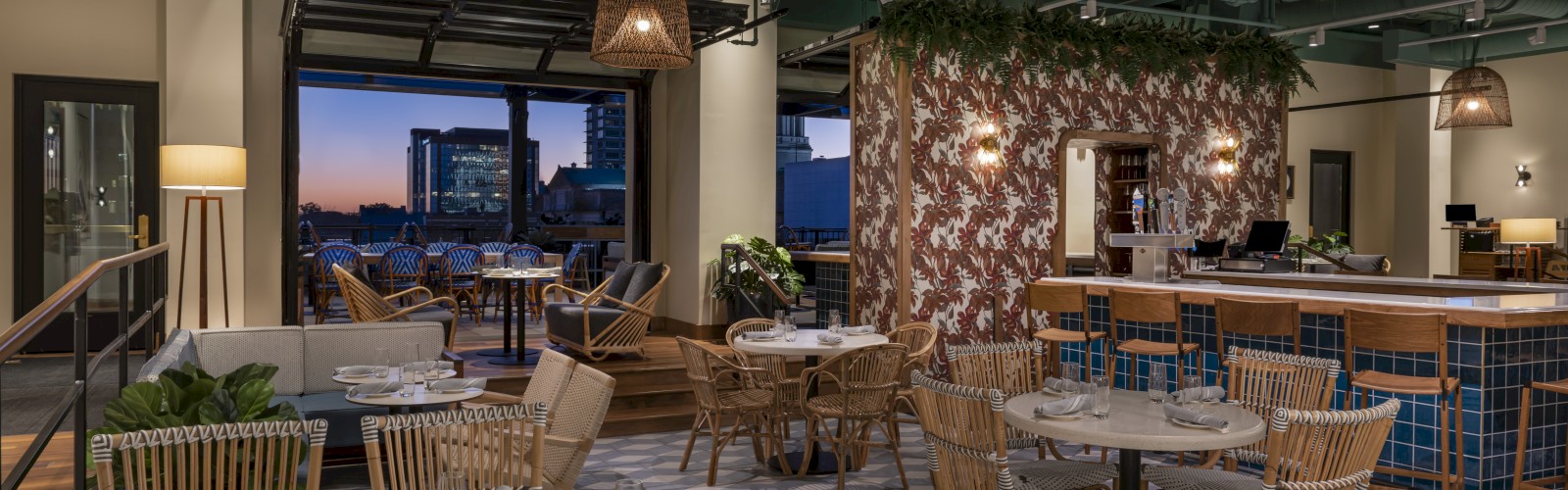 The image shows a cozy indoor restaurant with modern decor, wicker chairs, a bar, plants, and a partially open wall revealing a city view at dusk.