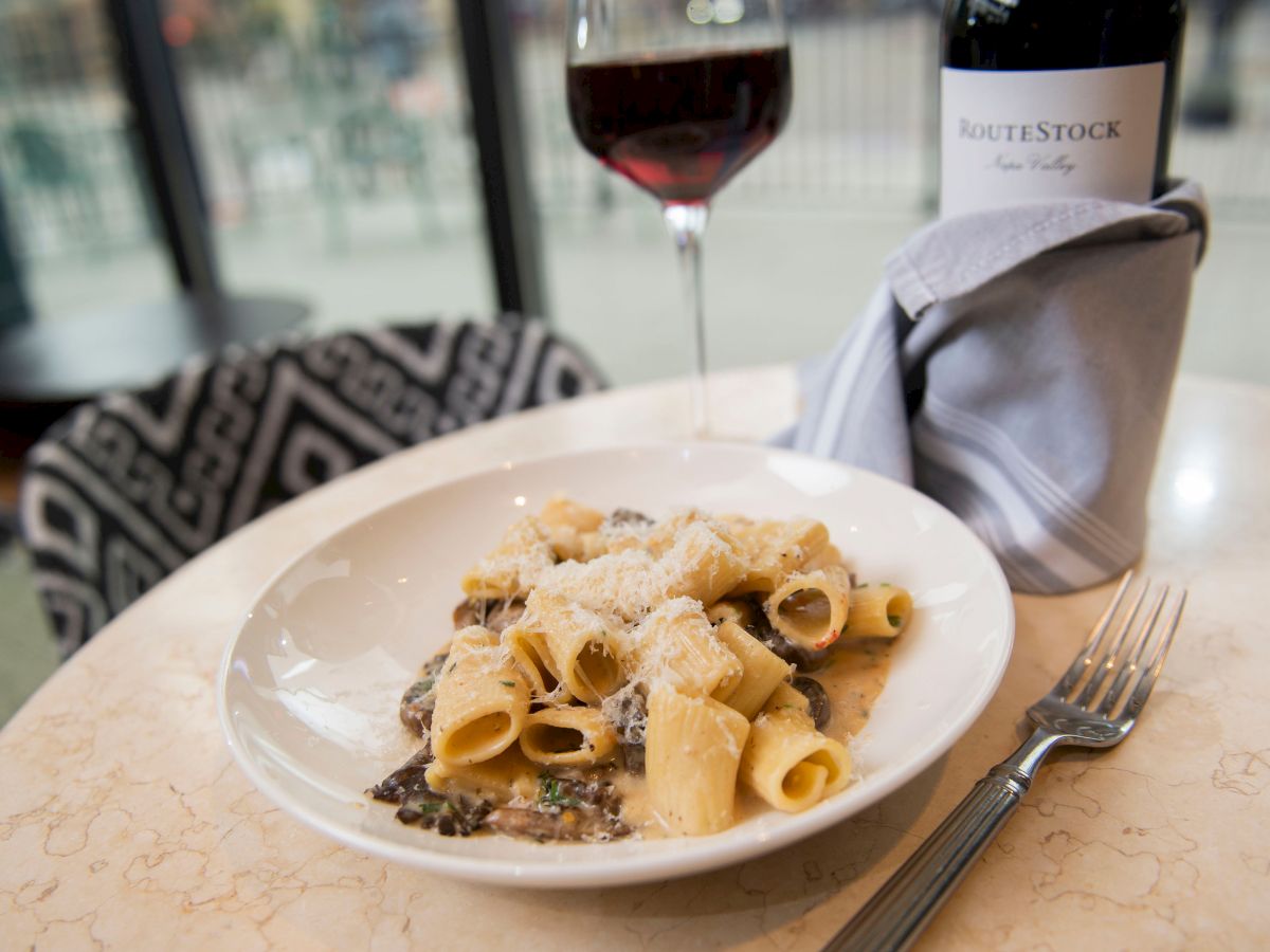 A pasta dish topped with cheese, a fork and knife on the side, a glass of red wine, and a bottle labeled 