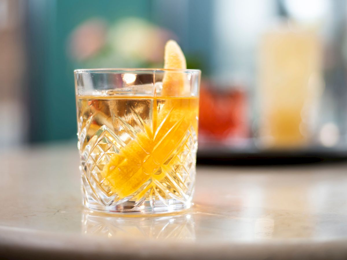 A crystal glass filled with a light brown drink, garnished with a lemon twist, sits on a round table in a softly blurred background.