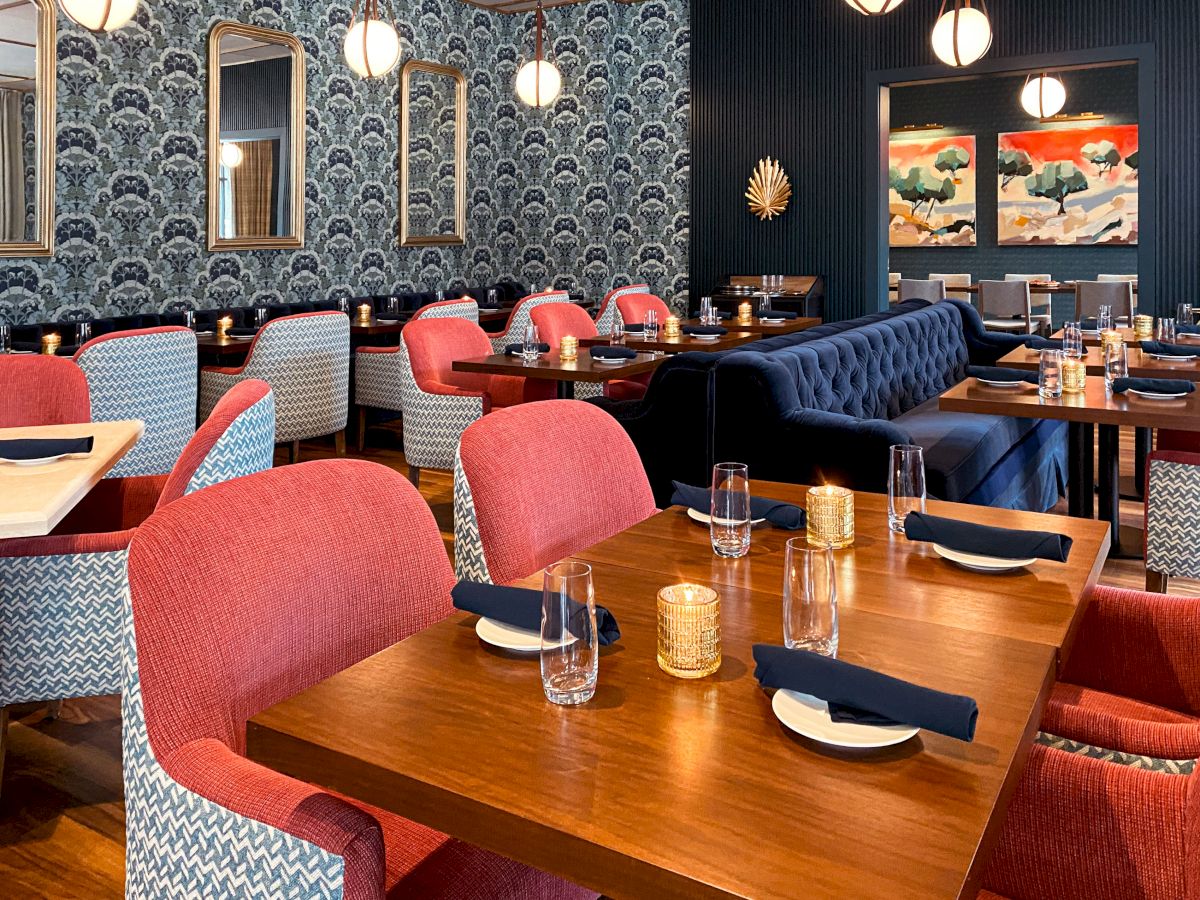 A stylish restaurant with patterned wallpaper, colorful chairs, wooden tables, and wall art, set with glasses and napkins.
