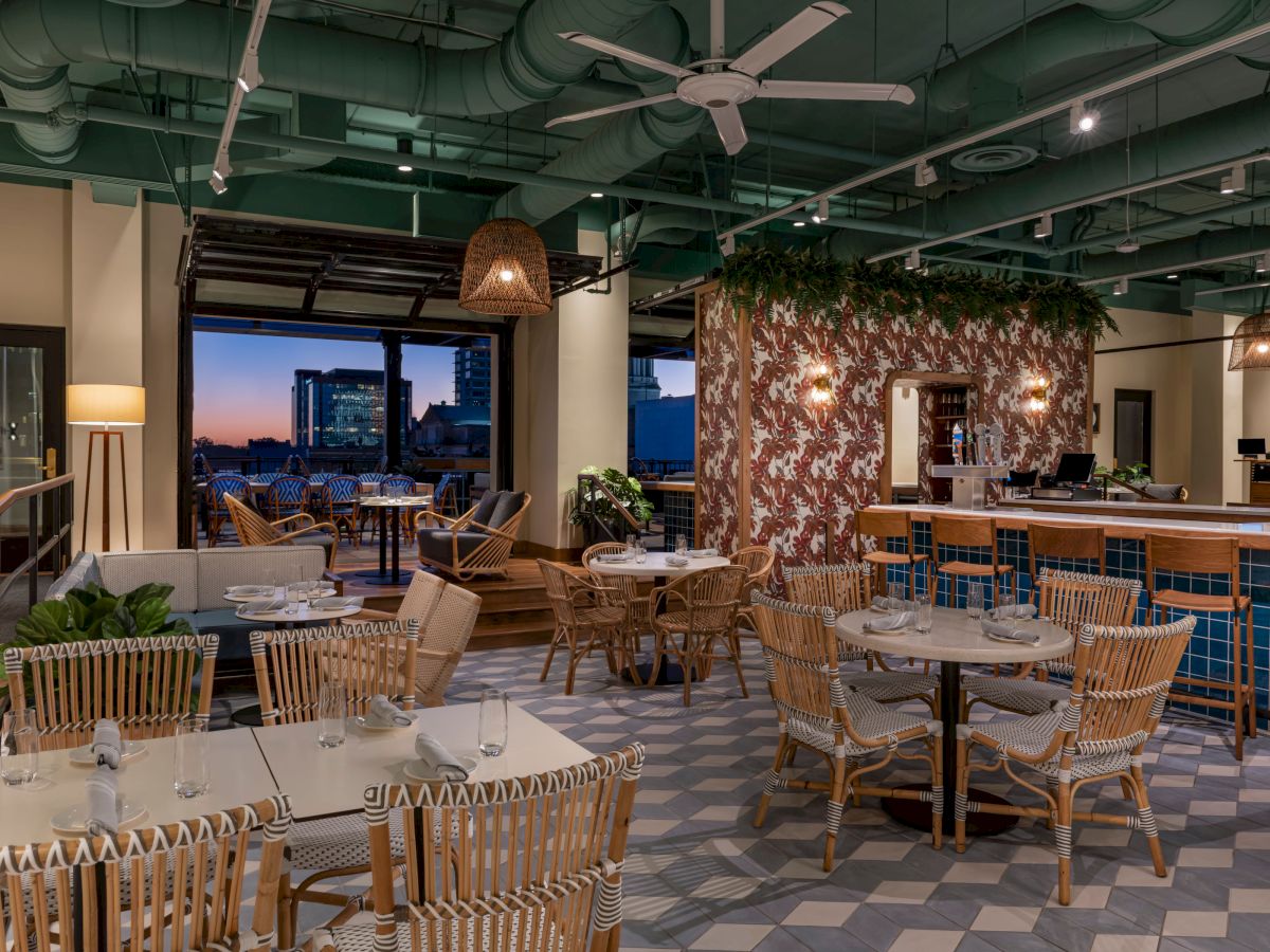 A modern, chic restaurant with wicker seating, a patterned floor, hanging lights, and a view of the city skyline through open windows.