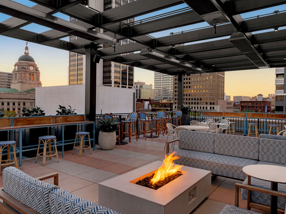 An outdoor rooftop seating area with fire pit, modern furniture, and city skyline views featuring a prominent domed building in the background.