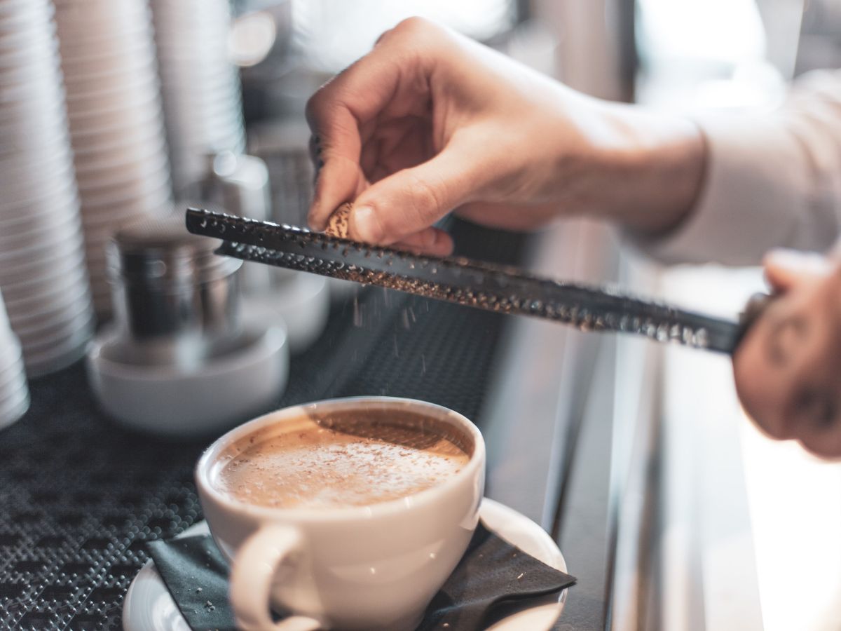 A person is grating something over a cup of coffee on a saucer with a dark napkin at a coffee station, with cups and other items in the background.