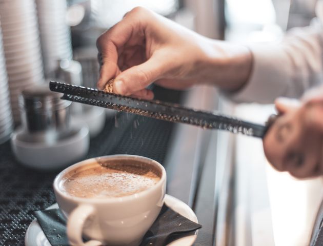 A hand grating something over a cup of coffee with foam in a white cup on a saucer, near coffee cups in a café setting.