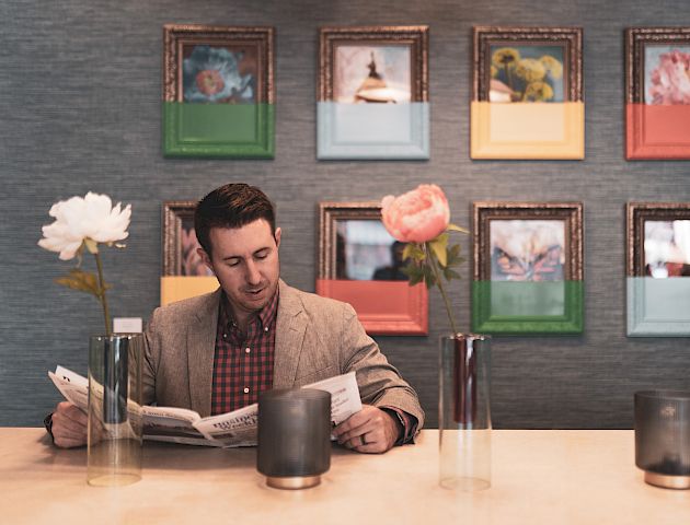 A man reading a newspaper at a table with vases of flowers, against a wall decorated with framed art.