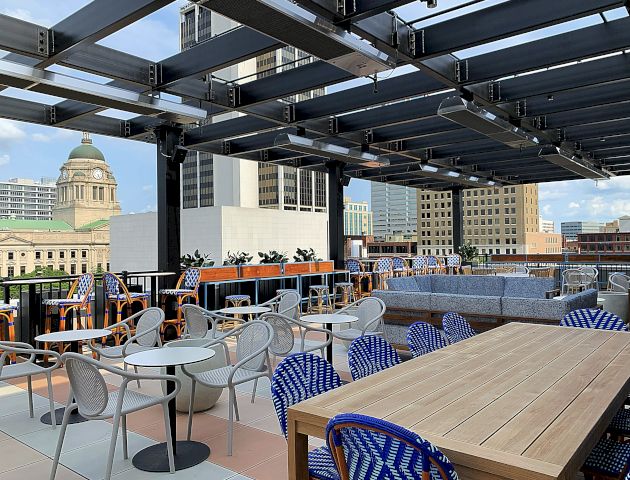 The image shows a rooftop patio with tables, chairs, and a cushioned seating area, overlooking a cityscape that includes a domed building.