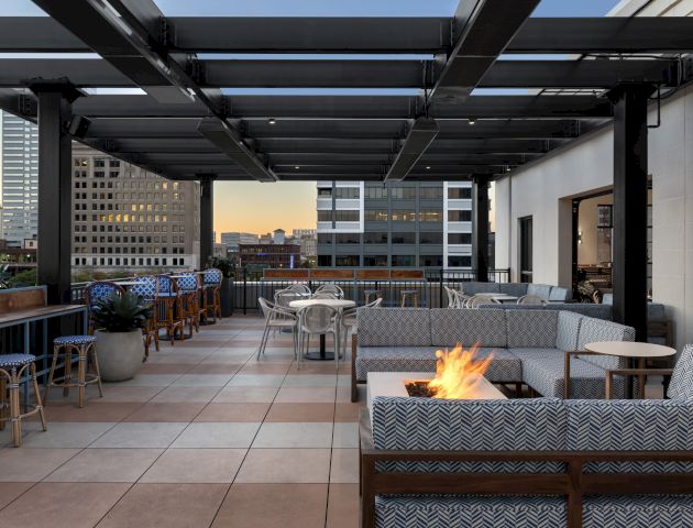 The image shows a modern rooftop patio with seating, tables, a fire pit, and city buildings in the background at sunset.