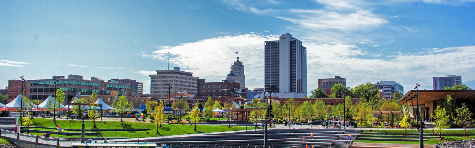 The image depicts a cityscape featuring green spaces, tall buildings, a calm body of water in the foreground, and a clear blue sky with clouds.