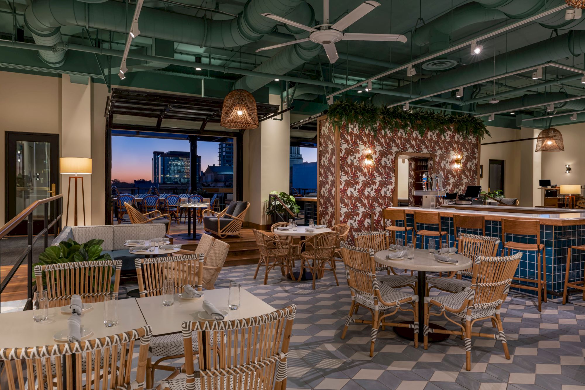 The image shows a cozy restaurant interior with wicker chairs, hanging light fixtures, and an open view of a cityscape at dusk.