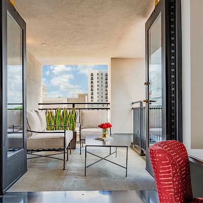 A modern balcony with seating, greenery, a coffee table with flowers, and a city view under a partly cloudy sky.