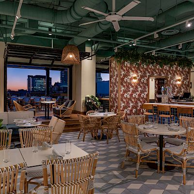 The image shows a stylish, modern restaurant with wicker furniture, large windows, plants, and a cozy ambiance under green ceiling ducts and fans.