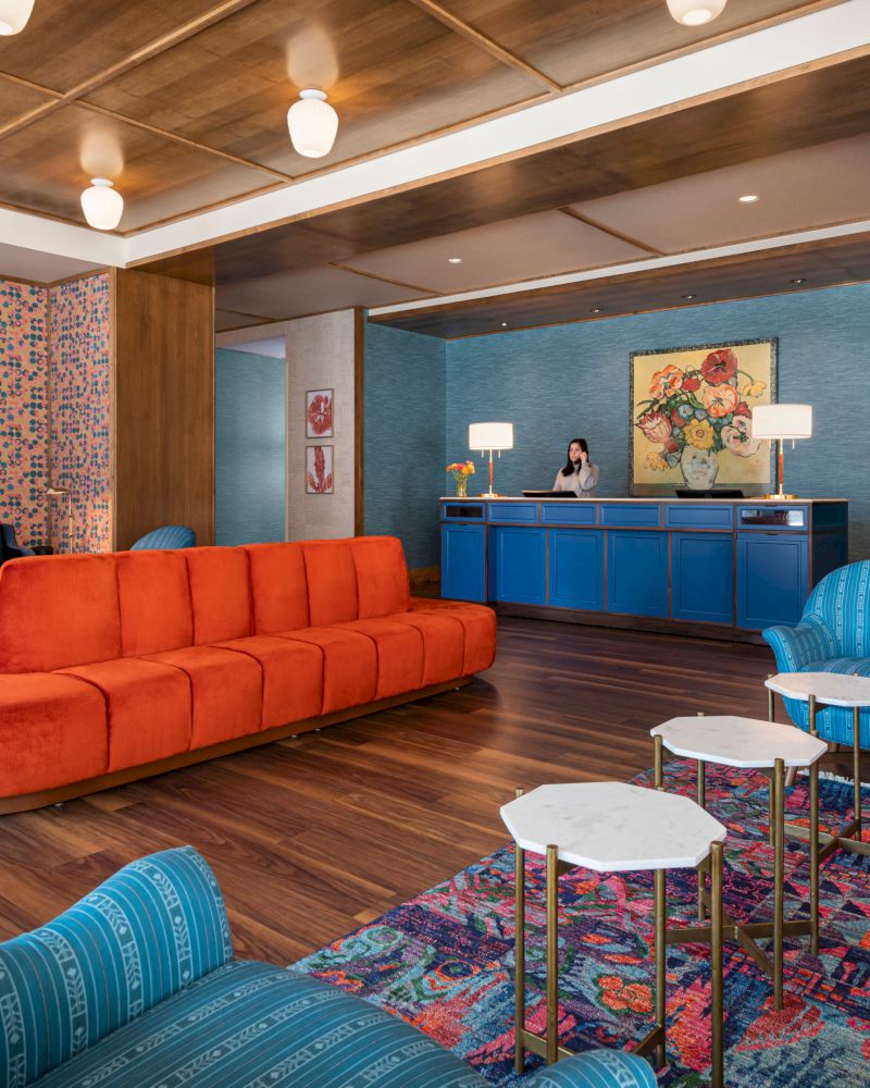 A modern hotel lobby with colorful furniture: a long orange sofa, blue chairs, white tables, and a reception desk adorned with lamps and artwork.