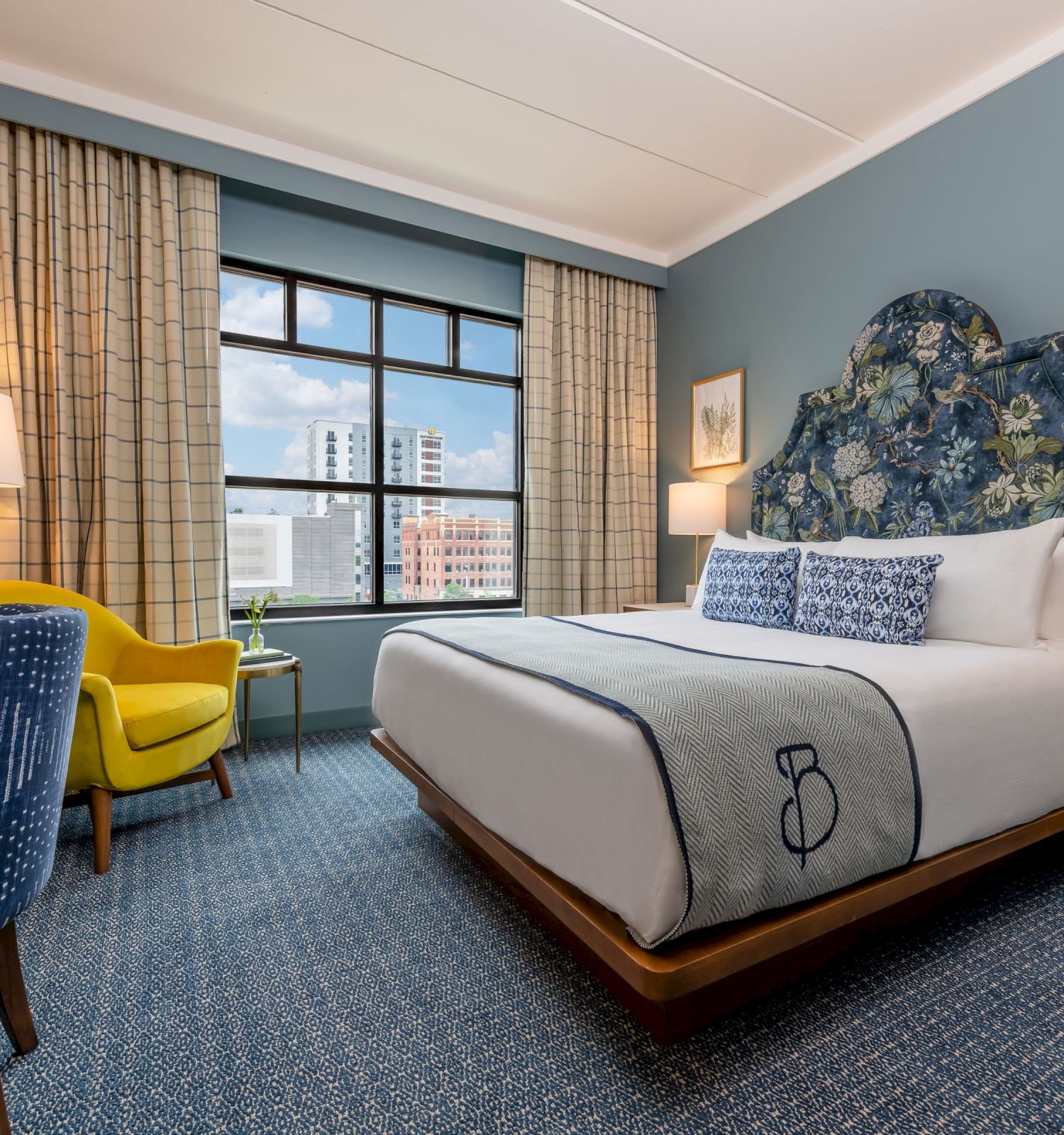 A cozy hotel room with a king-size bed, a desk, a yellow chair, and a window with curtains showcasing a city view. Decor includes lamps and wall art.