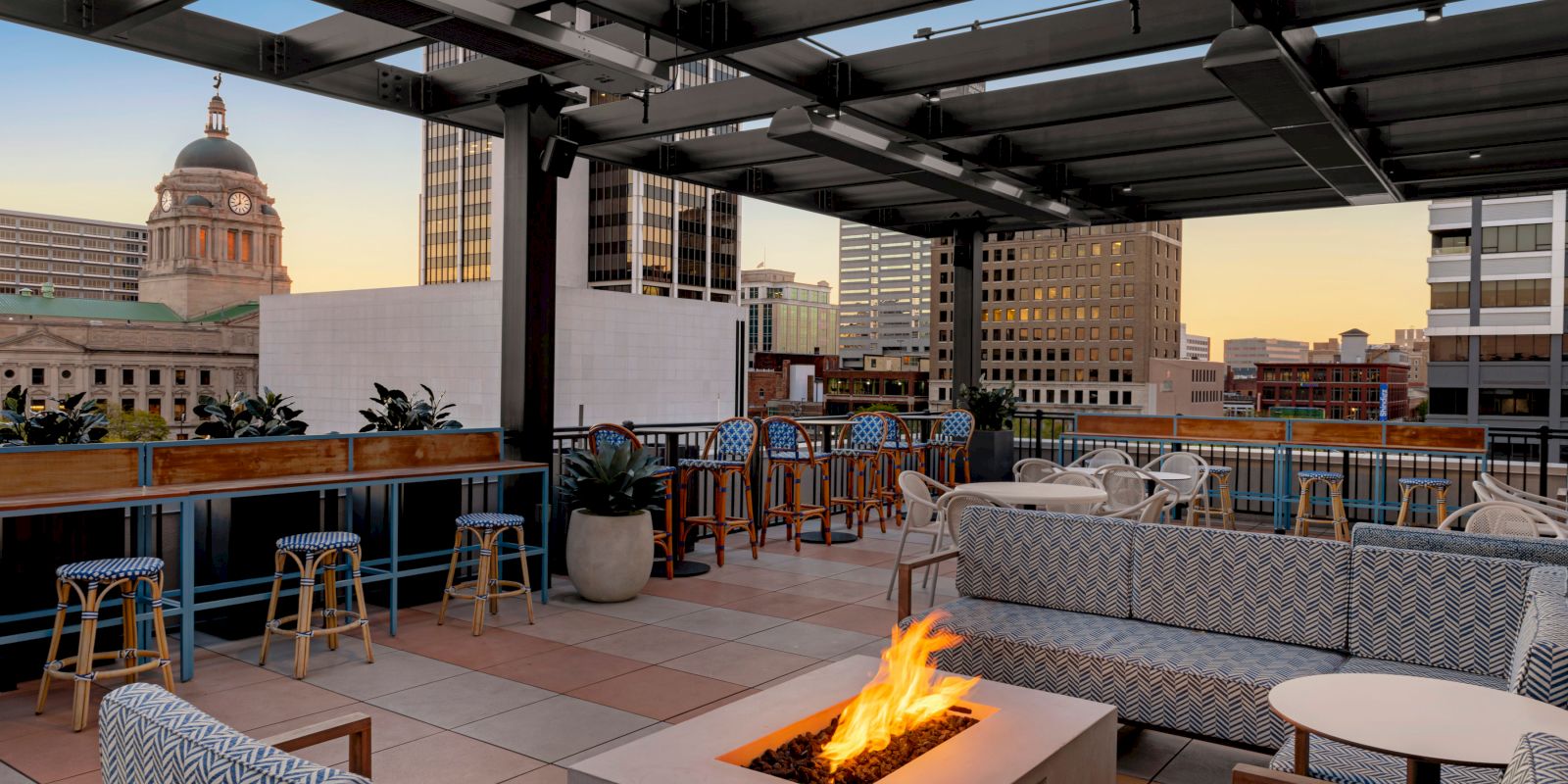 A rooftop lounge with modern seating, a fire pit, bar stools, and cityscape views during sunset. Tall buildings and a dome structure are visible.