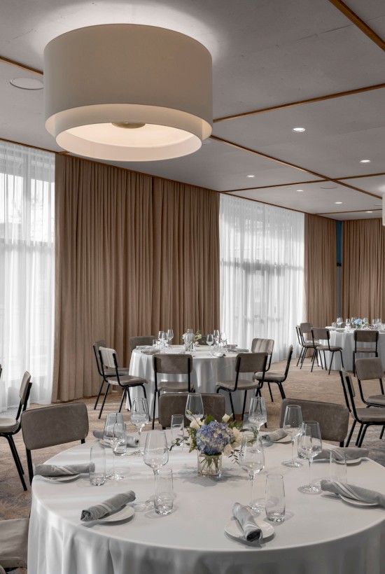 The image shows a banquet hall set up for an event, with round tables covered in white tablecloths, chairs, and elegant place settings.