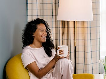 A person with curly hair is sitting on a yellow chair, holding a mug, and smiling. There is a lamp and a window with checked curtains behind them.