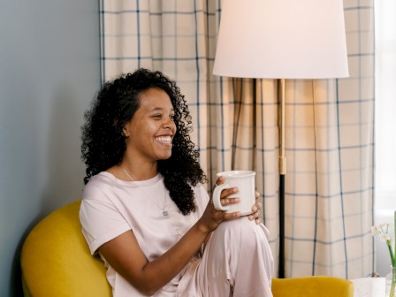 A person with curly hair is sitting on a yellow chair, holding a mug, and smiling. There is a lamp and a window with checked curtains behind them.