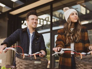 Two people with bicycles standing outdoors, one wearing a striped coat and beanie, the other in a dark jacket, both smiling.
