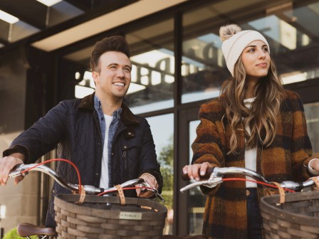 Two people with bicycles standing outdoors, one wearing a striped coat and beanie, the other in a dark jacket, both smiling.