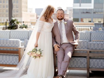 A bride and groom pose for a photo on an outdoor patio, with the bride in a white gown holding a bouquet and the groom in a light-colored suit.