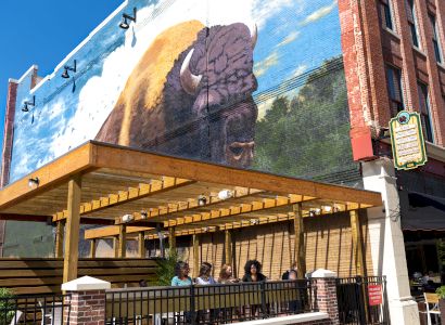 A group of people sits under a wooden pergola outside a brick building with a large mural of a bison on the wall.