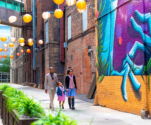 A family of three walks past colorful graffiti and hanging lanterns in an alley, surrounded by brick walls and greenery.