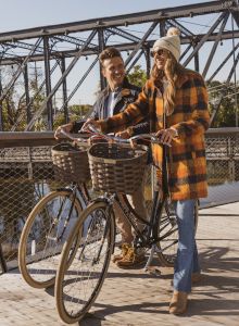 Two people walk with bicycles on a wooden walkway by a bridge. One wears a checkered coat and beanie, the other a jacket.