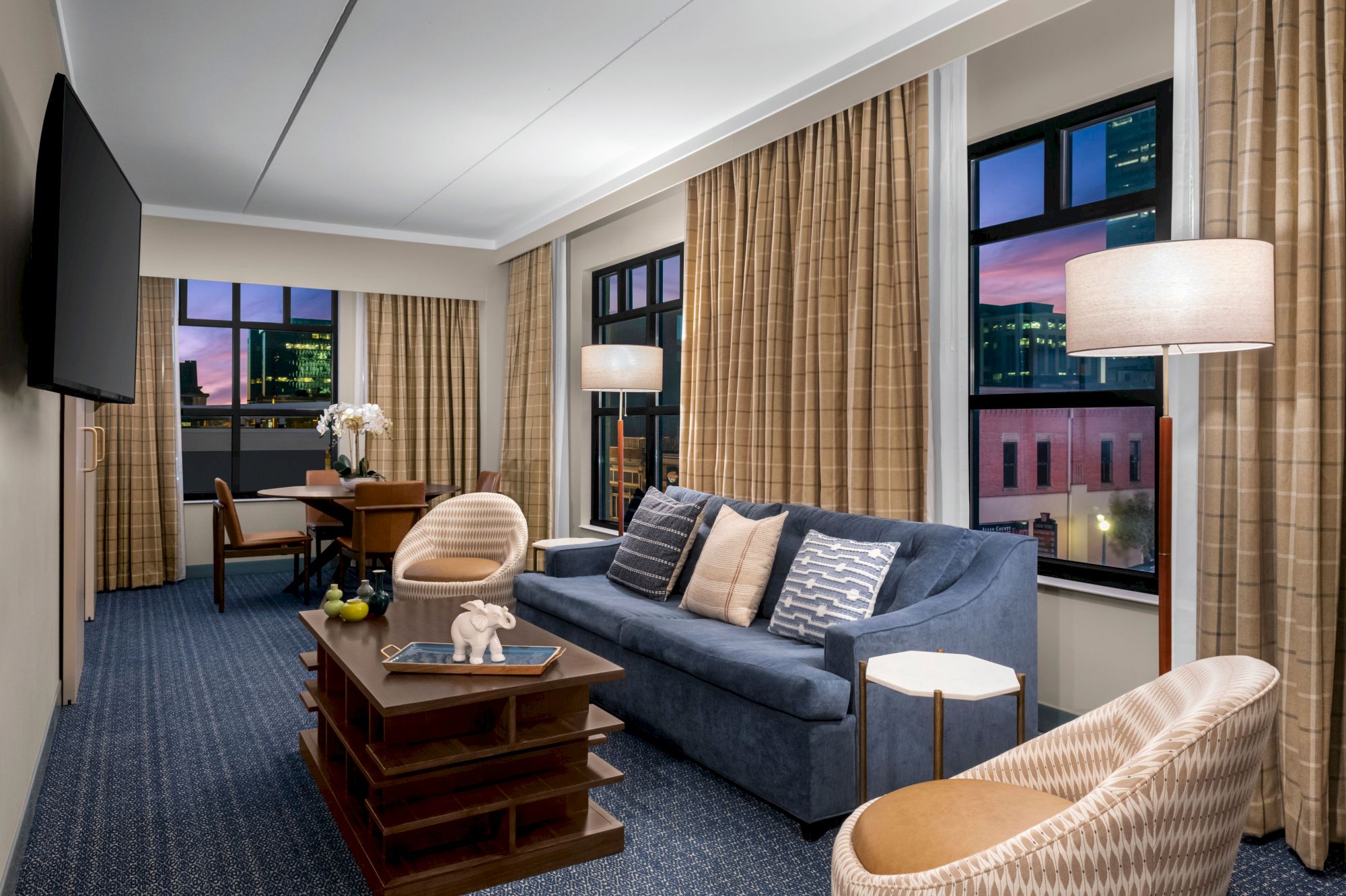 This image shows a modern hotel room with a seating area, featuring a blue sofa, armchairs, coffee table, floor lamps, and large windows with city views.