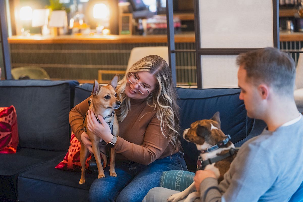 Two people sitting on a couch, each holding a small dog, and the person on the left smiling at their dog, indoors.