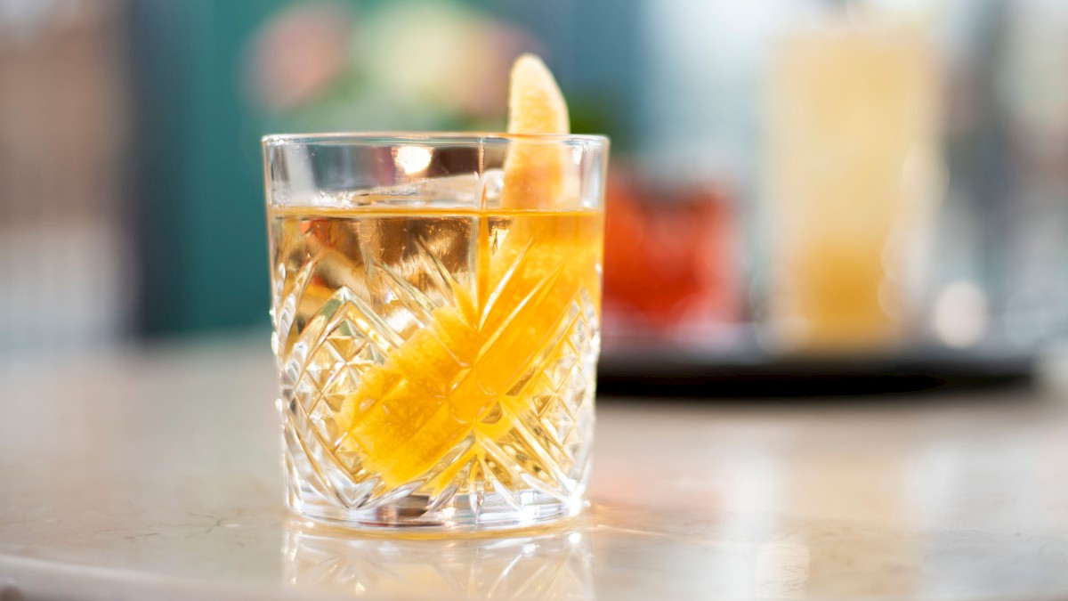 The image shows a crystal glass with a cocktail garnished with a lemon twist, placed on a marble table.