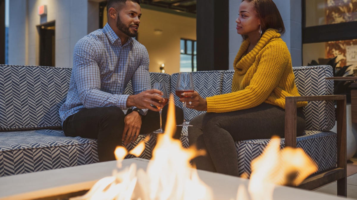 A man and a woman are seated on an outdoor sofa, holding wine glasses, with a firepit in the foreground, sharing a conversation.