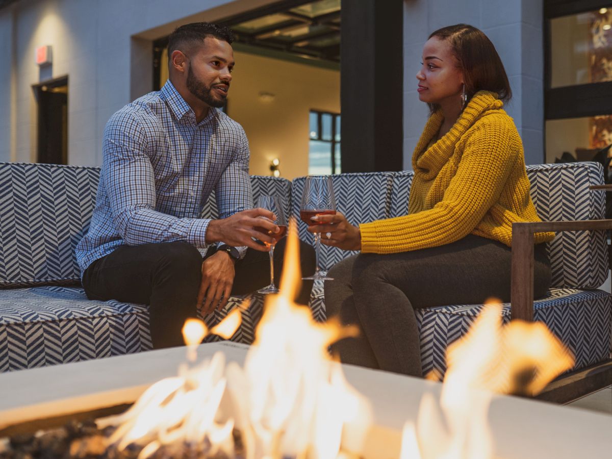 A man and a woman are seated on an outdoor sofa, holding wine glasses, with a firepit in the foreground, sharing a conversation.