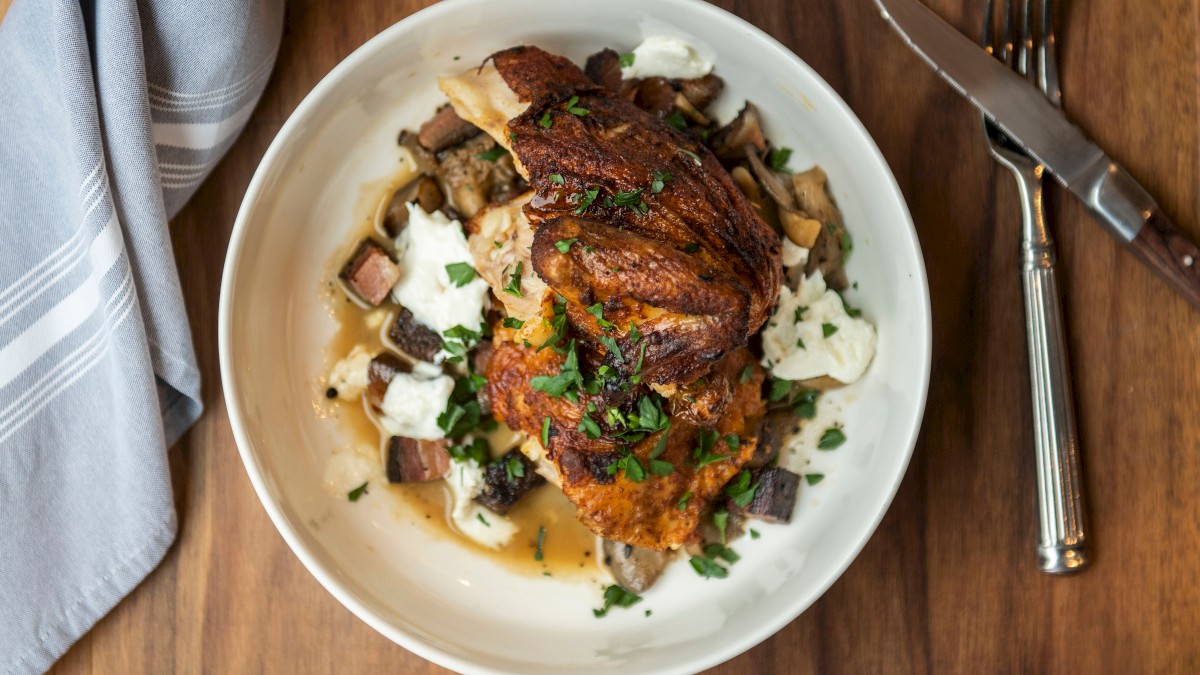 A plate of roasted chicken with herbs served over a bed of vegetables and garnished with fresh parsley, placed on a wooden table with utensils.