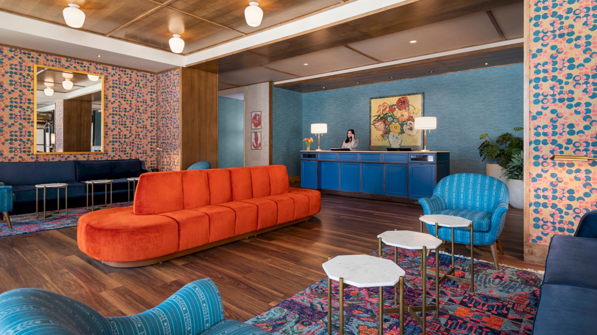 A modern hotel lobby with vibrant, colorful furniture, including an orange sofa, blue armchairs, patterned wallpaper, and a reception desk in the background.