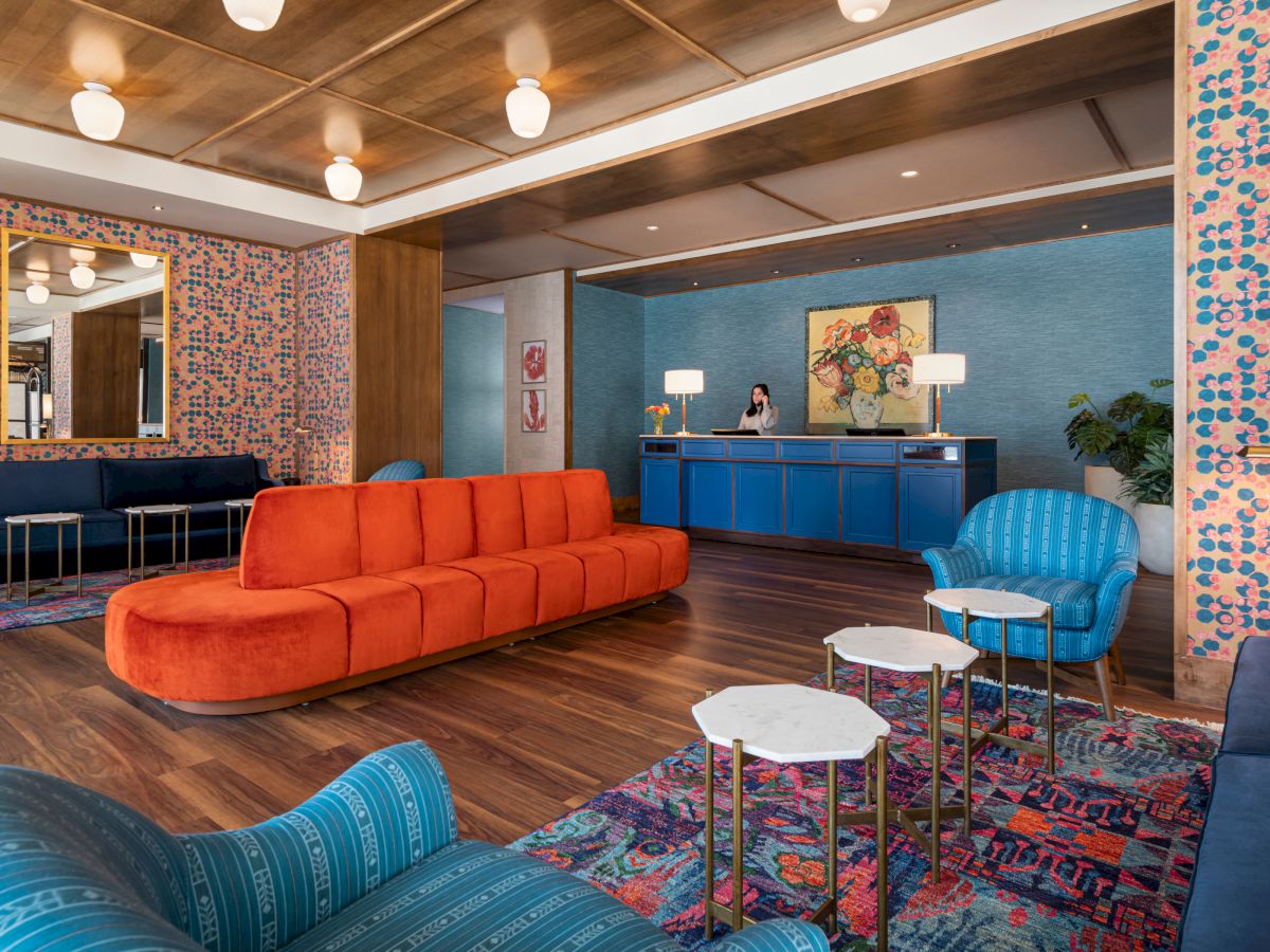 A modern hotel lobby with vibrant, colorful furniture, including an orange sofa, blue armchairs, patterned wallpaper, and a reception desk in the background.