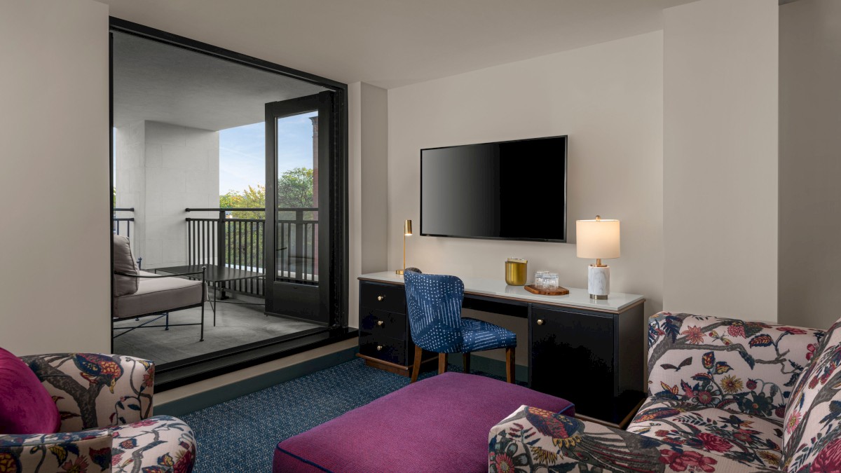 The image shows a cozy living area with floral armchairs, a purple ottoman, a desk with a chair, a table lamp, a wall-mounted TV, and a balcony.