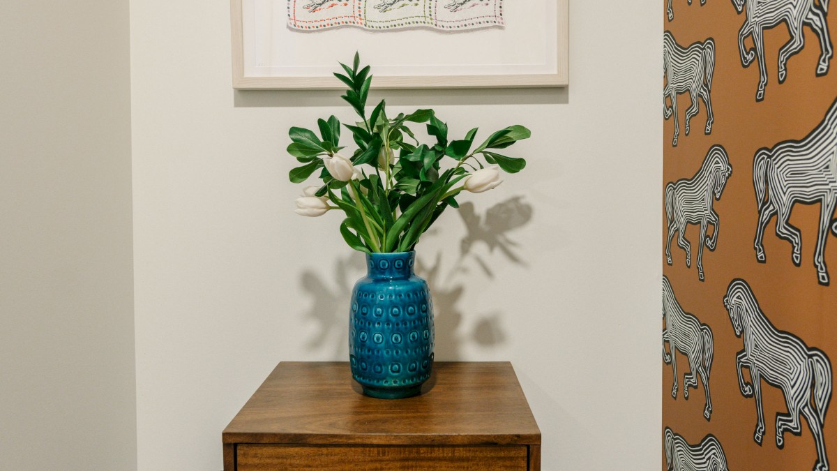 A small side table with a blue vase and green plant, artwork above, and wallpaper with zebra patterns on the right wall in a corner space.