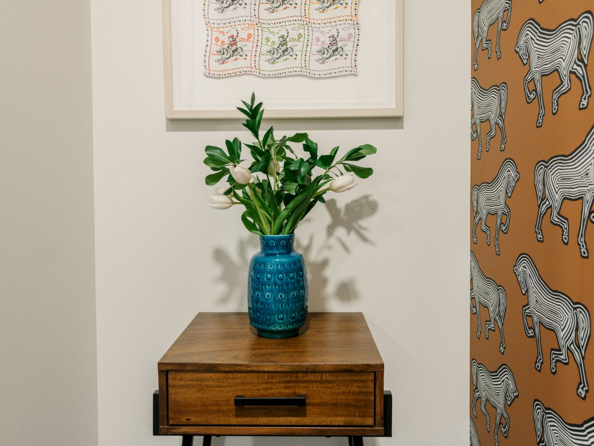 A small side table with a blue vase and green plant, artwork above, and wallpaper with zebra patterns on the right wall in a corner space.