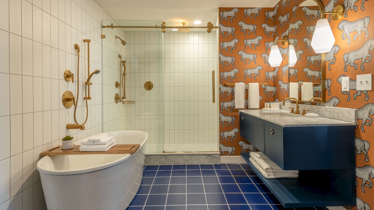 A modern bathroom with blue tile floor, freestanding tub, brass fixtures, glass-enclosed shower, animal-patterned wallpaper, and double sink vanity.