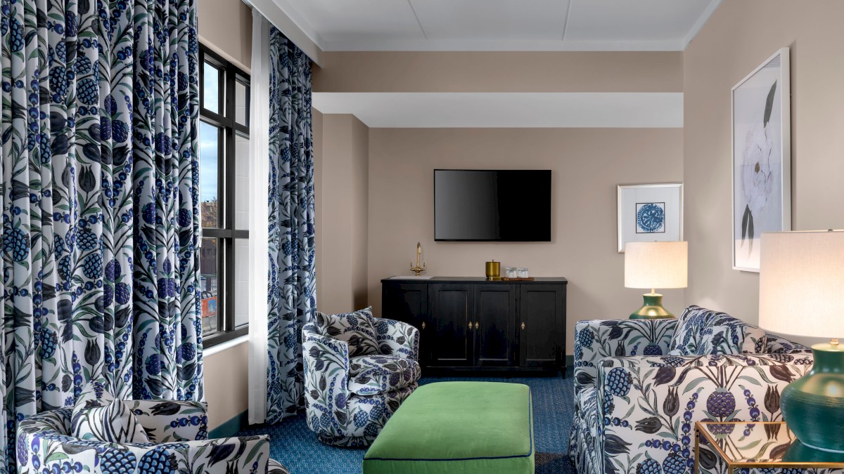 A stylish living room with floral-patterned curtains and furniture, green ottoman, TV, cabinet, table lamp, and wall art on beige walls.