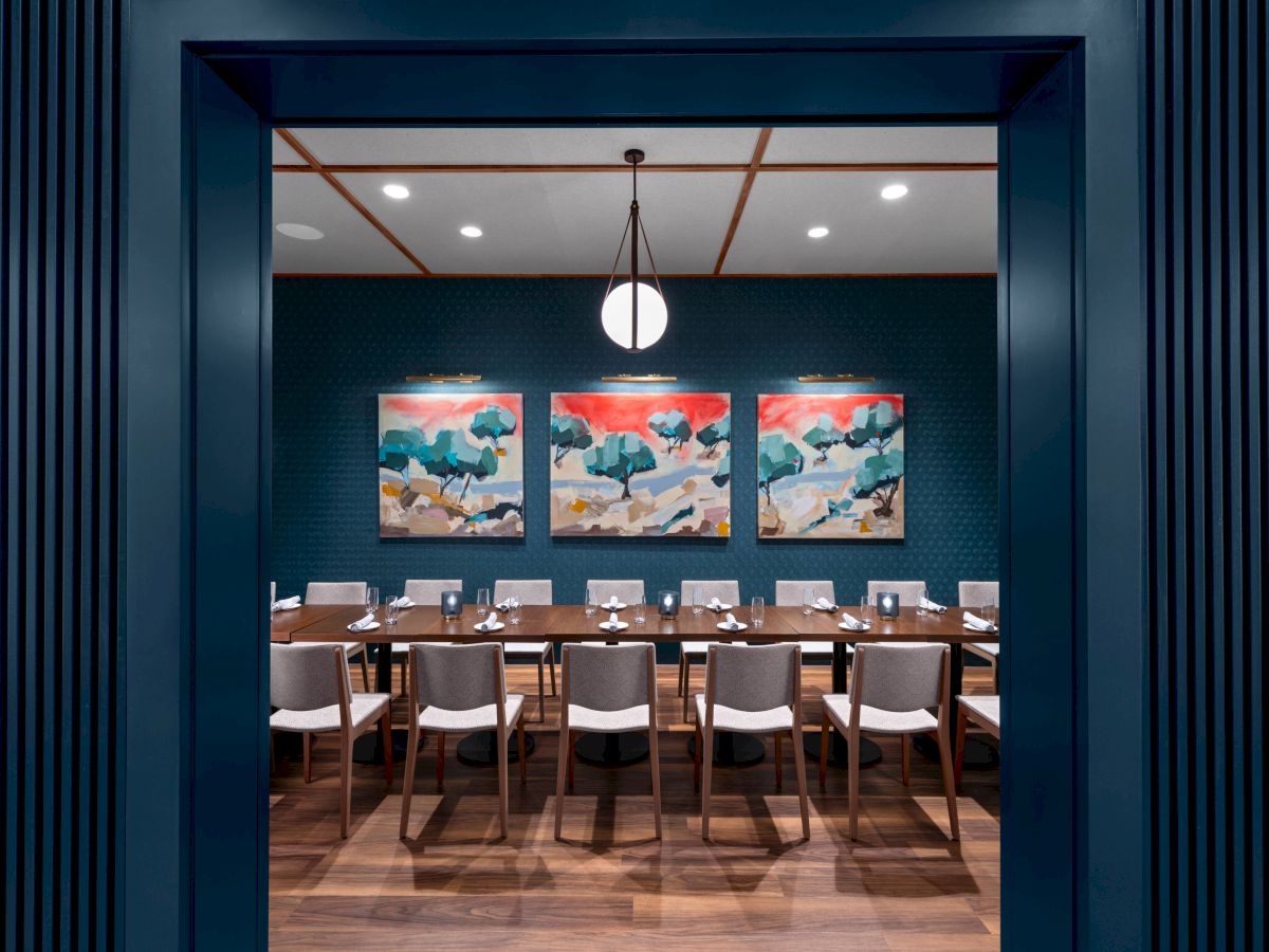 The image shows a long dining table with chairs in a modern room, featuring three colorful abstract paintings on the back wall and a hanging light fixture.