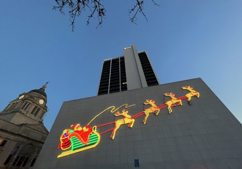 A building features a large, illuminated display of Santa in a sled pulled by reindeer, under evening sky with a historic structure nearby.