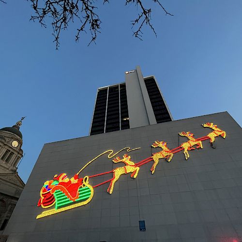 A building features a large, illuminated display of Santa in a sled pulled by reindeer, under evening sky with a historic structure nearby.