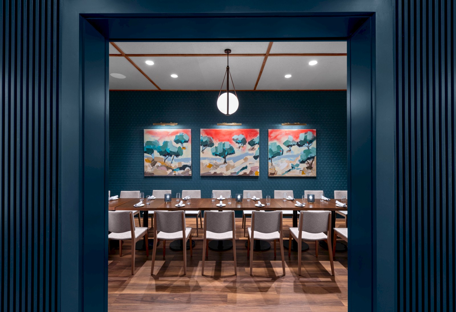 A modern dining room with a long table, multiple chairs, three colorful paintings on the wall, and a single round ceiling light.