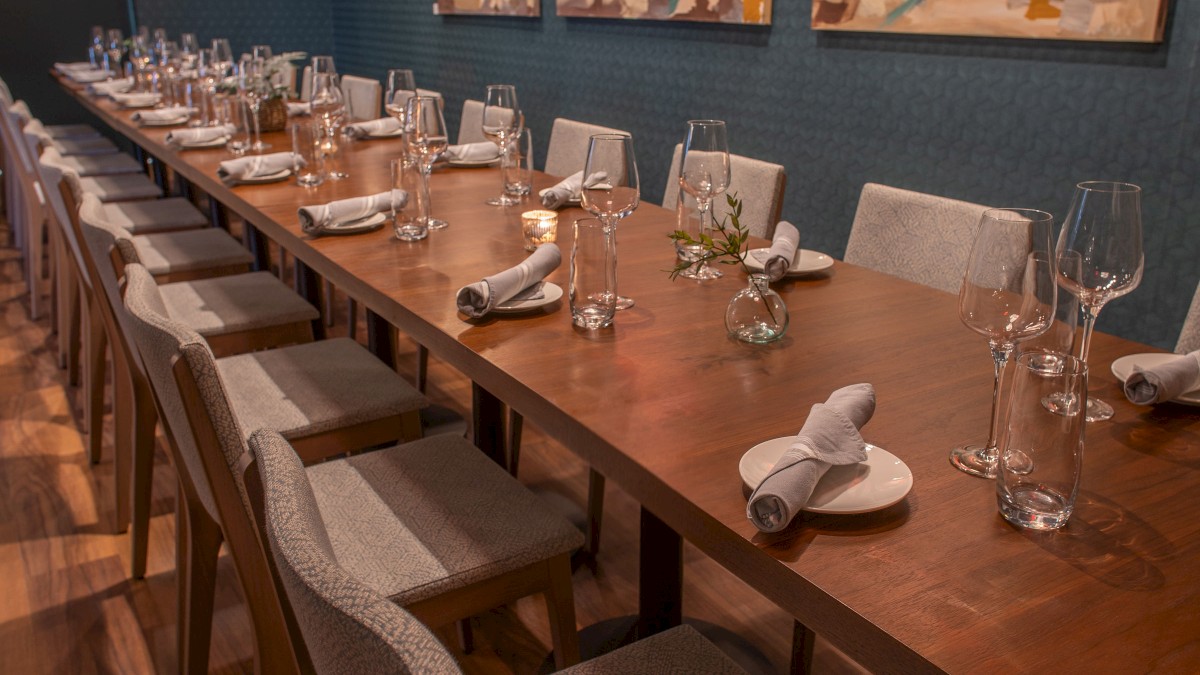 A dining room with a long wooden table, set with glasses, napkins, and cutlery. The walls feature two colorful paintings under warm lighting.