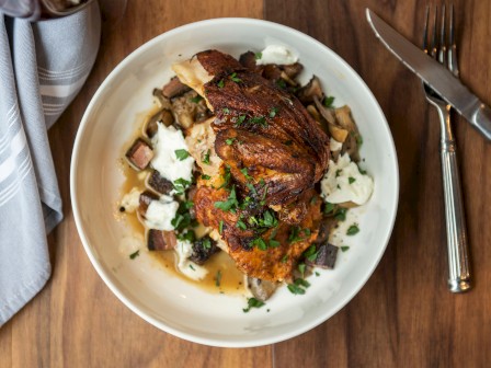 A roasted chicken dish garnished with parsley, resting on a bed of vegetables and creamy sauce, placed on a wooden table with cutlery and a napkin.