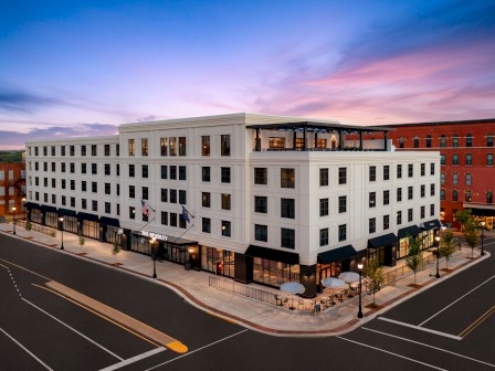 A modern, multi-story building on a street corner at dusk, featuring ground-floor shops, outdoor seating, and flagpoles by the entrance.