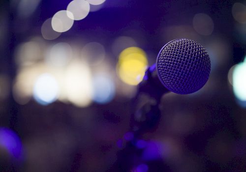 The image shows a close-up of a microphone with a blurred, colorful background, likely capturing a stage or performance setting.