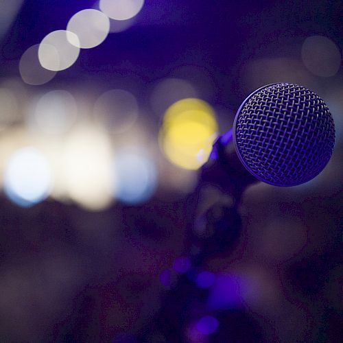 The image shows a close-up of a microphone with a blurred, colorful background, likely capturing a stage or performance setting.