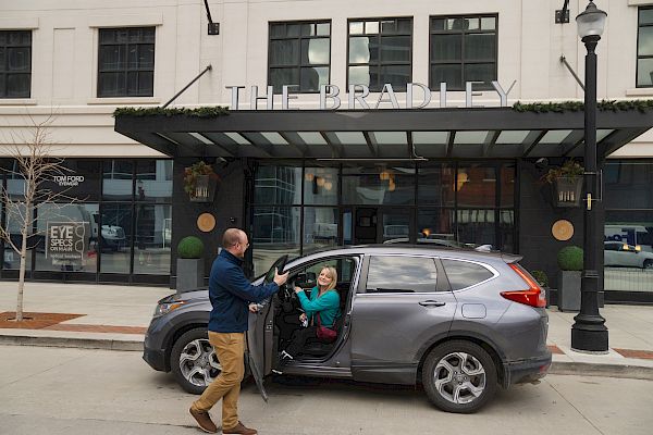 A man is helping a woman exit a gray SUV parked in front of a building with a sign reading 